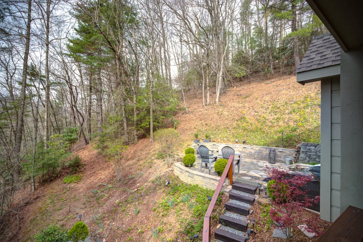 A stone seating area bordered by wooden steps and lush garden plants sits in a tranquil, wooded setting. The natural landscape invites guests and their pets to enjoy the outdoors, whether playing, relaxing, or socializing around the fire pit.