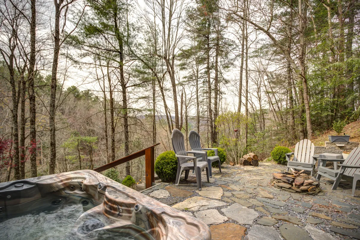 The image depicts a cozy stone patio with a hot tub and Adirondack chairs, set against a wooded backdrop. The relaxed scene is ideal for unwinding in the hot tub or gathering outdoors for stargazing and stories.
