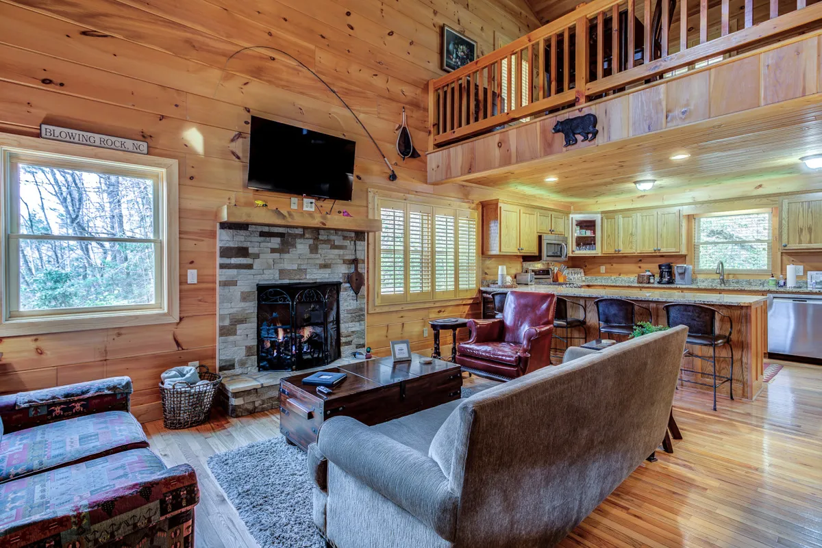 This image captures the open-concept living area, featuring a cozy sofa, a prominent fireplace, a spacious kitchen island, and large windows letting in forest views. The setup encourages socializing, relaxation, and shared activities.