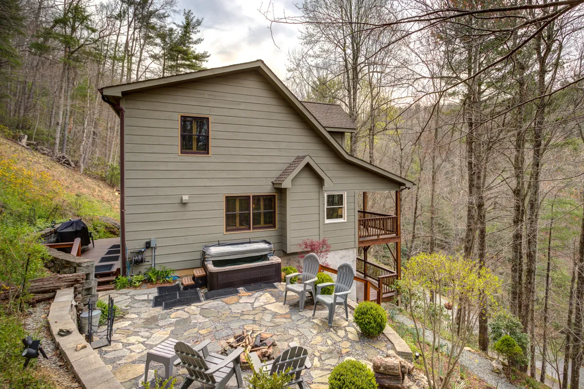 The exterior image showcases a spacious stone patio nestled among lush trees, featuring a bubbling hot tub, a glowing fire pit surrounded by Adirondack chairs, a dining table for outdoor meals, and beautifully landscaped gardens. The setting feels private and tranquil, inviting guests to relax and socialize in a serene mountain atmosphere.