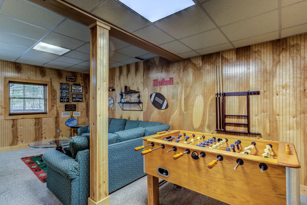 A lively game room setting featuring a foosball table as the focal point, surrounded by a green couch and wood-paneled walls. Playful decorative signs add to the energetic, welcoming vibe, and the layout suggests ample room for friends or family to gather and socialize.