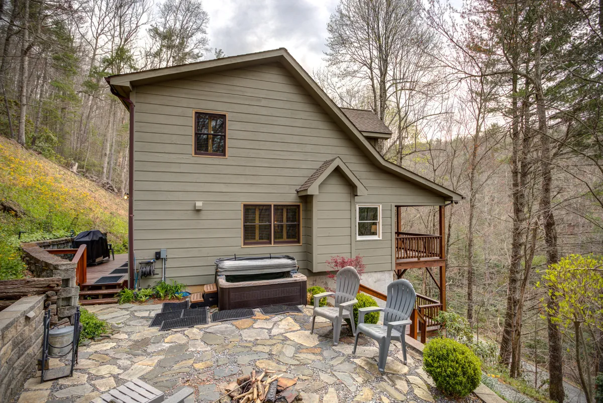 This image shows a spacious wooden deck nestled among tall trees, featuring a private hot tub and a stone patio. The area is surrounded by lush greenery, providing a tranquil setting for relaxation or gatherings. The hot tub is prominently placed for guests to enjoy the beautiful forest views.
