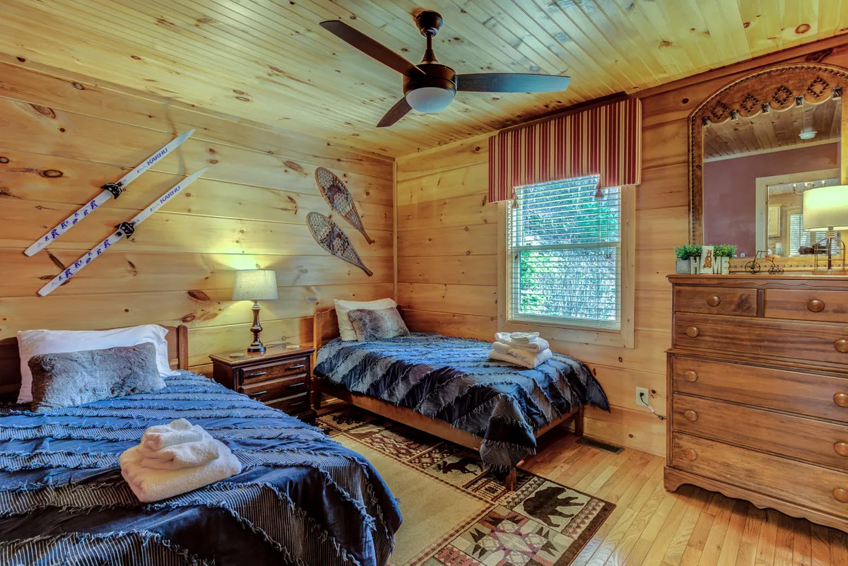 A cozy loft space with two twin beds dressed in blue bedding, a wooden dresser, a lamp, and playful ski-themed wall decor. The arrangement suggests a cheerful and inviting spot for kids or extra guests to relax, rest, or play board games.