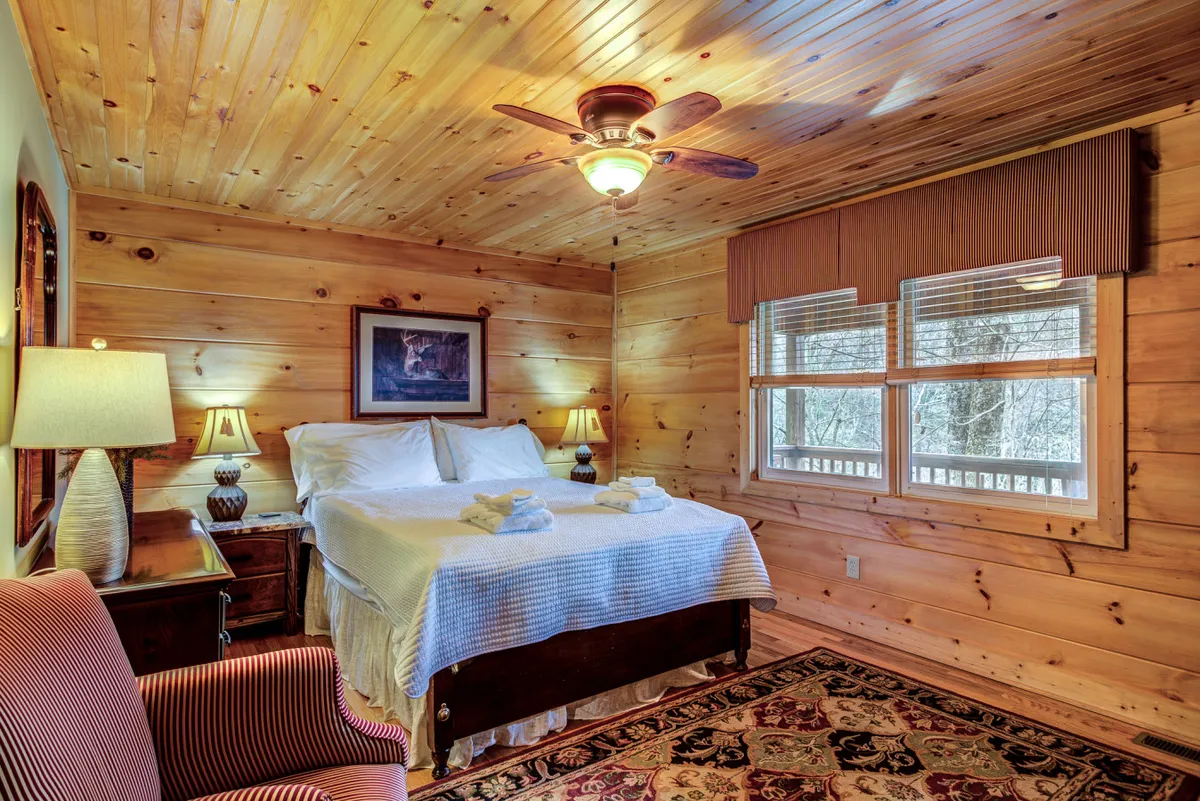 A serene bedroom with a plush bed, an armchair by the window, nightstand lamps, a rug, and a large window letting in morning light. The space feels comfortable and tranquil—ideal for unwinding or reading.