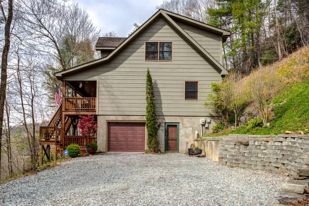A view of the front of the cabin with a welcoming porch, large windows, and the garage door. The scene is framed by lush greenery, suggesting an environment ideal for family gatherings and relaxing outdoors. The spacious porch hints at morning coffees or evening downtime after a day exploring local attractions.