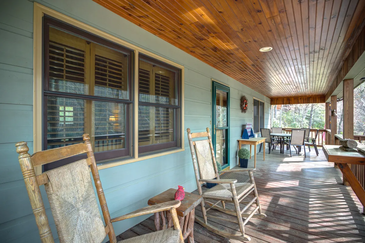 This exterior image shows a welcoming porch with wooden rocking chairs, a dining table, and a cozy green door under a wood-paneled ceiling. The scene is warm and inviting, suggesting a perfect spot for groups to gather and share meals outdoors or enjoy the mountain sunset together.
