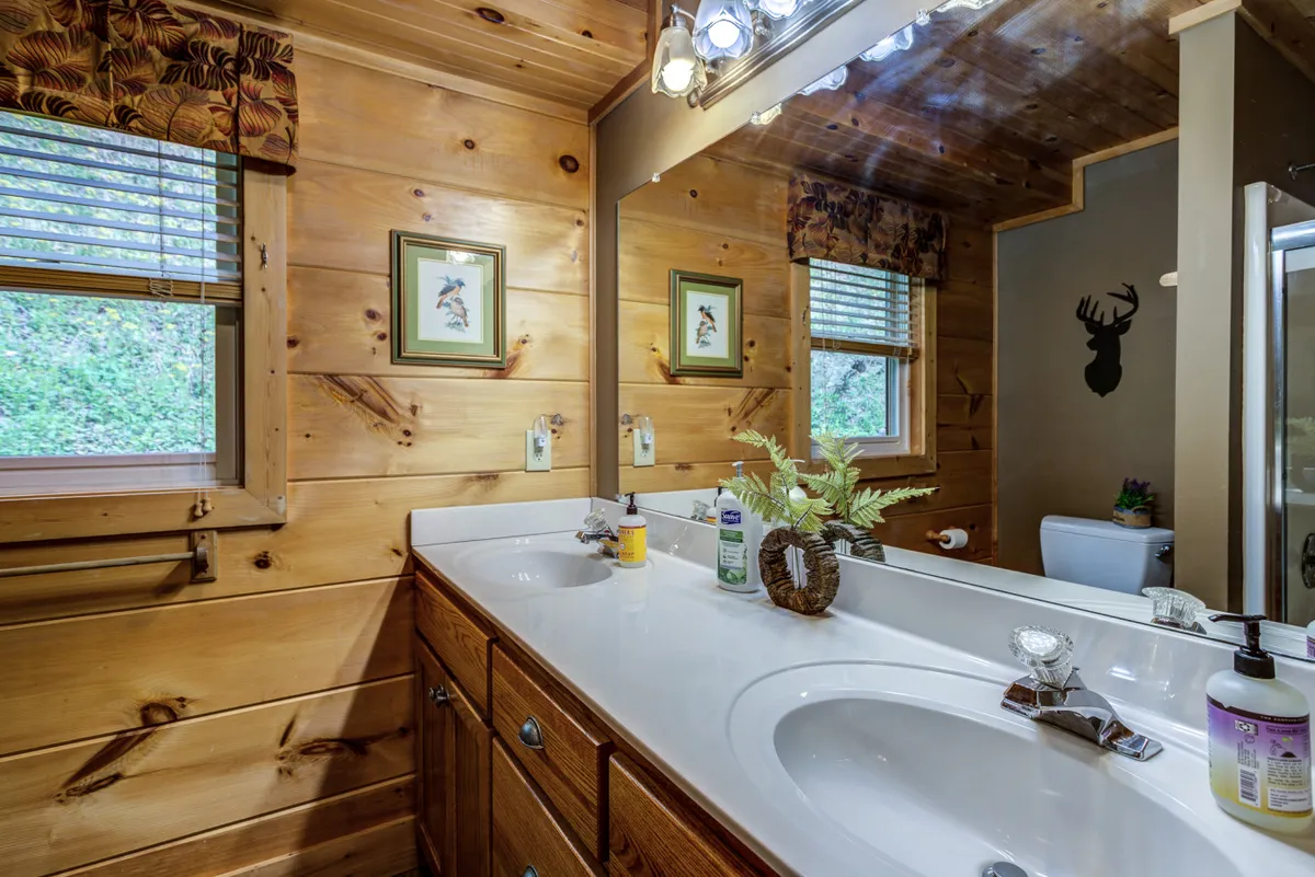 A warmly lit bathroom with a double-sink vanity, wooden cabinetry, decorative wall art, and a window looking out onto greenery. The space is both functional and relaxing, ideal for group privacy and convenience.